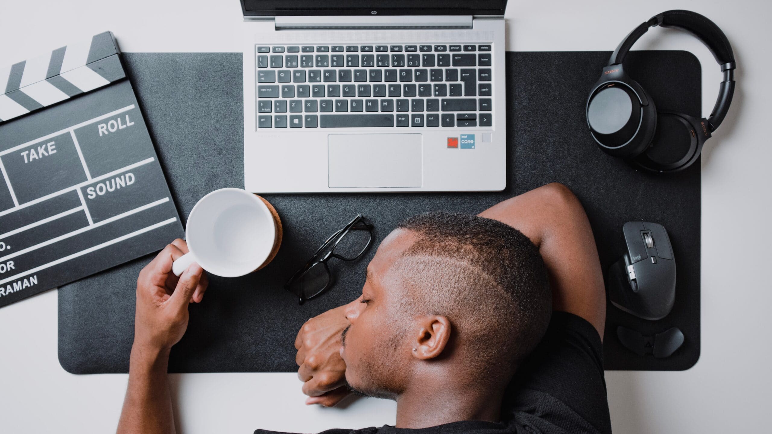 man sleeping on top of his desk. on the deks: laptop, headphones, mouse, glasses and cup.