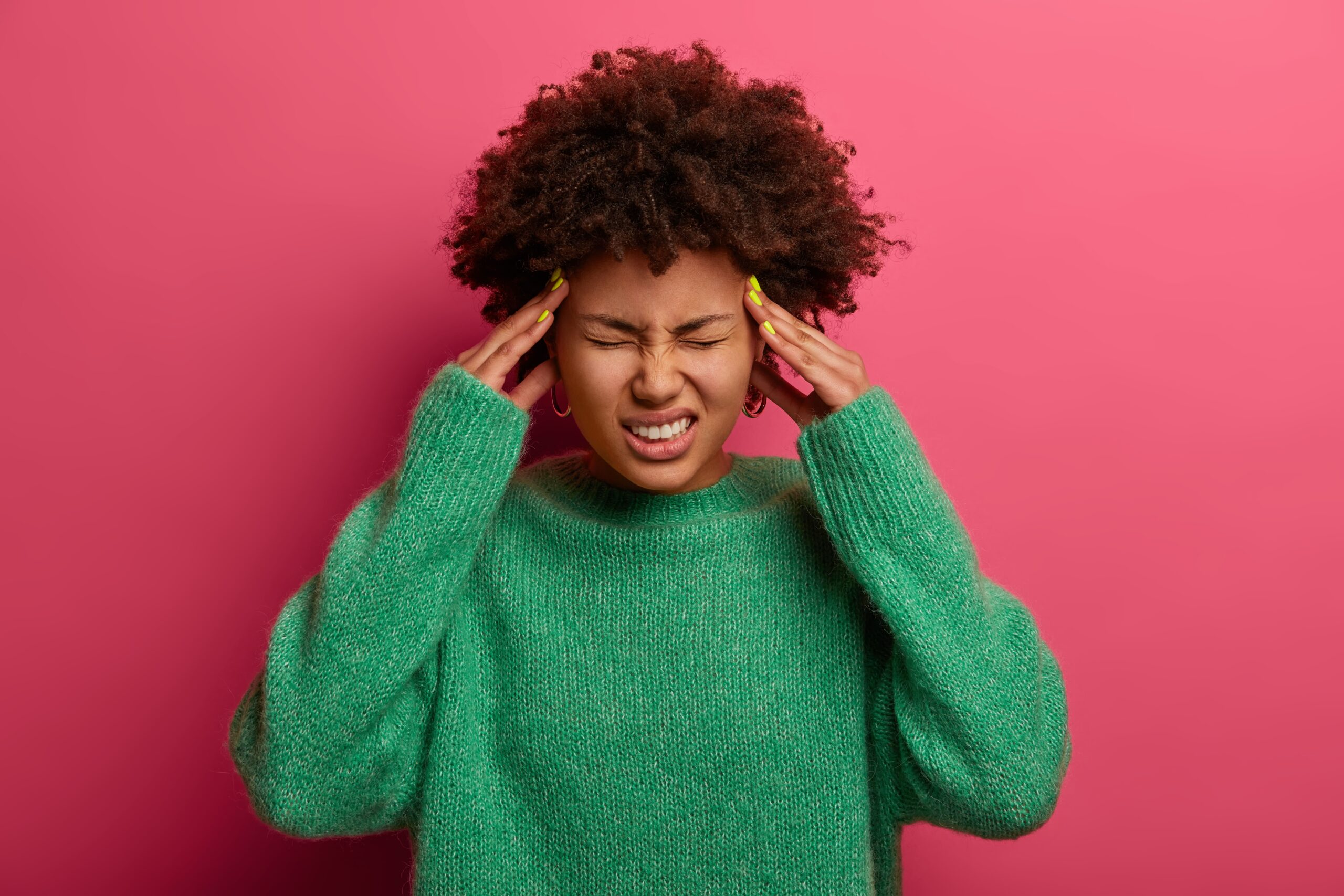 Upset dark skinned curly woman touches temples, frowns face and suffers from migraine, terrible headache, needs painkillers, wears warm green sweater, closes eyes to relieve pain, poses indoor